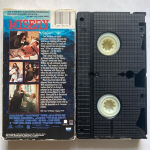 May include: A VHS video cassette of the film "Misery", with the title in red. The cover shows scenes from the movie and text detailing the plot. The cassette is black with two circular reels.