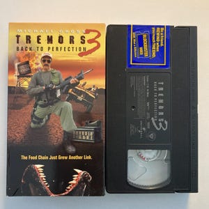 May include: A VHS tape and its cover for the movie "Tremors 3: Back to Perfection." The cover features a man holding a gun, with the film's title and a tagline. The VHS tape has a Blockbuster Video sticker.