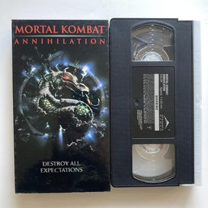 May include: A Mortal Kombat Annihilation VHS tape and its black cardboard sleeve. The sleeve features a green dragon graphic, blue light effects, and the text Mortal Kombat Annihilation Destroy All Expectations. The black tape has a label with text.