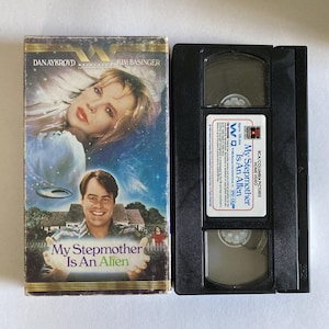 May include: A VHS tape and case for the movie "My Stepmother Is An Alien." The case features images of Dan Aykroyd and Kim Basinger, with the title prominently displayed. The black VHS tape has a white label with the movie title.