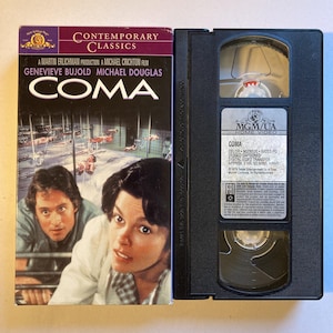 Peut inclure: Une cassette VHS du film "COMA" avec Genevieve Bujold et Michael Douglas sur la jaquette. La cassette noire porte une étiquette avec le titre du film et d'autres informations. Le logo MGM/UA Home Video est également visible.