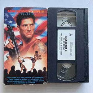 May include: A VHS tape and its cardboard sleeve for the movie Kickboxer 3: The Art of War. The sleeve features a man holding a firearm, two fighters in a ring, and an American flag background. The black plastic tape has a white label with text.