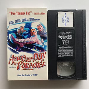 May include: A VHS tape of the film Another Day in Paradise, featuring a white cardboard sleeve with a movie scene of people in a car and the title in red and yellow text. The black plastic cassette has a white label reading Gone Hollywood Video.