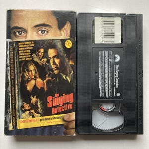 May include: A VHS tape and its case for "The Singing Detective" movie. The case features images of the cast, including Robert Downey Jr. The tape is black with a white label and a barcode.