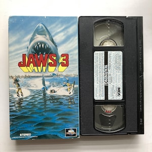 May include: A Jaws 3 VHS tape and its cardboard sleeve. The sleeve features a large shark emerging from blue water with water skiers and the text Jaws 3. The black plastic tape has a white label with the title Jaws 3, warning text, and MCA Home Video branding.