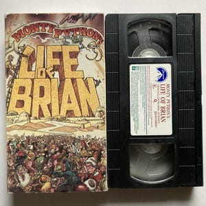 May include: A vintage VHS tape of Monty Python's "Life of Brian" with its original cover art. The cover features a crowd scene and the film's title in large, bold letters. The tape itself is black with a white label.