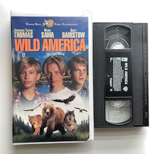 May include: A Wild America VHS tape and its plastic case. The cover features actors Jonathan Taylor Thomas, Devon Sawa, and Scott Bairstow, with a mountain landscape, a bear, a wolf, a cougar, and birds. The black tape has a white label.