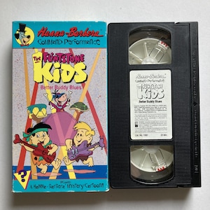 Peut inclure: Une cassette VHS et son étui pour "The Flintstone Kids: Better Buddy Blues". L'étui présente des personnages de dessins animés jouant d'instruments, avec du texte incluant "Hanna-Barbera" et "COMMAND Performance".