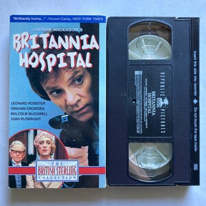 May include: A VHS video cassette and its cover for the film "Britannia Hospital." The cover displays the film's title and a close-up of a person's face. The tape is black with white text and a clear window to view the tape.