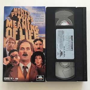 May include: A VHS tape case for the Monty Python's The Meaning of Life film. The case is black with a red and white label featuring the film title and a group of actors from the film.