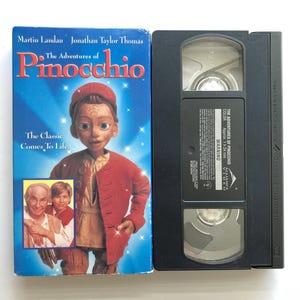 May include: A VHS tape of The Adventures of Pinocchio featuring Martin Landau and Jonathan Taylor Thomas. The blue cover displays a wooden puppet character in a red jacket, with a small inset photo of the actors. A black VHS cassette is included.