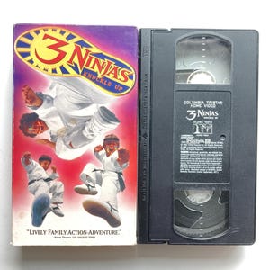May include: A 3 Ninjas Knuckle Up VHS tape and its cardboard sleeve. The sleeve features three children in white martial arts uniforms against a purple and red background with the movie title in yellow and blue. The black plastic tape has a label.