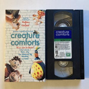 May include: A VHS tape and its cover for the animated series "Creature Comforts." The cover features cartoon characters and the text "Creature Comforts." The VHS tape is black with a blue and green label.
