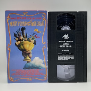 May include: A VHS tape and its cover for the movie "Monty Python and the Holy Grail." The cover is blue with a gold border and features a cartoon illustration of the film's characters. The VHS tape is black with white text.