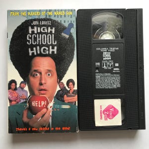 May include: A VHS tape case for the comedy film "High School High" starring Jon Lovitz. The cover features a man with an afro holding an apple with the word "HELP!" on it. The tagline reads "There's a new teacha in the Hood!"