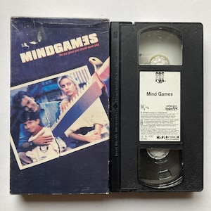 May include: A VHS tape and case for the movie "Mind Games". The case is dark blue with the movie title in white and red. The VHS tape is black with a white label that reads "Mind Games".