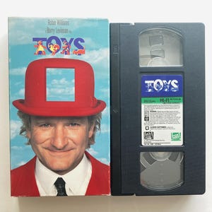 May include: A VHS tape of the film Toys starring Robin Williams. The cover features a man in a red suit and red bowler hat with a square cutout showing a blue sky. The black plastic cassette has a blue label with the title Toys and text.