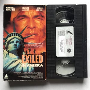 May include: A VHS tape and its cover for the movie "Exiled in America." The cover features a dramatic image of a man's face, the Statue of Liberty, and the film's title. The tape is black with a white label.
