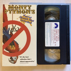 May include: A vintage VHS tape and its cover for "Monty Python's Parrot Sketch Not Included." The cover features a red prohibition sign over a parrot illustration. The tape is black with a white label.