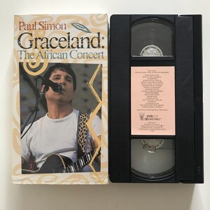 May include: A VHS tape and its cover for Paul Simon's "Graceland: The African Concert." The cover features a photo of Paul Simon singing and playing guitar. The tape is black with a pink label listing songs.