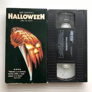 May include: A VHS tape of John Carpenter's Halloween featuring a black case with a graphic of a hand holding a knife against a carved pumpkin. The tape itself is black plastic with HGV branding and the title Halloween printed on the label.