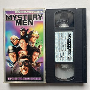 Peut inclure: Une cassette VHS du film "Mystery Men" avec une jaquette colorée représentant des personnages costumés. La cassette noire est à côté de la jaquette avec le texte "Special Edition" et "They're Not Your Average Superheroes".