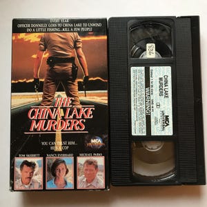 May include: VHS video cassette tape of the movie "The China Lake Murders". The cover features a man in a police uniform standing in a desert landscape. The text "Every year Officer Donnelly goes to China Lake to unwind. Do a little fishing...kill a few people." is at the top of the cover. The text "You can trust him...He's a cop" is below the title. The actors Tom Skerritt, Nancy Everhard, and Michael Parks are listed below the text.