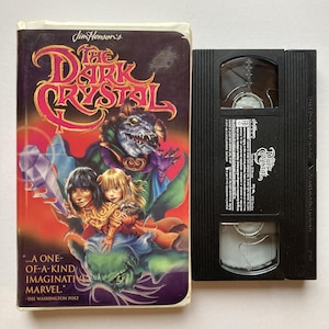 May include: A VHS tape and a book titled "The Dark Crystal." The book cover features a vibrant illustration with characters and the title in pink and gold. The VHS tape is black with the title on the label.