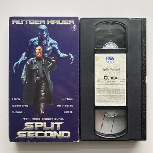 May include: A VHS tape and its cardboard sleeve for the film Split Second starring Rutger Hauer. The purple sleeve features a dark creature behind a man in a trench coat with the text: He's seen the future... Now he has to kill it.
