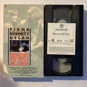 May include: A VHS tape and its cover for the movie "Hearts of Fire." The cover features Fiona, Everett, and Dylan, with the movie title in red. The VHS tape has a white label with the movie title and Warner Home Video branding.