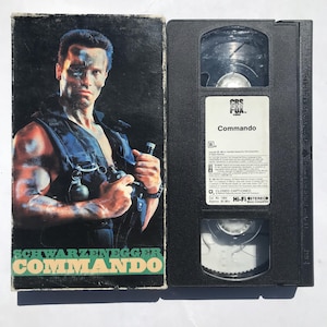 May include: A VHS tape case with a black and white image of Arnold Schwarzenegger on the front. The title of the movie is "Schwarzenegger Commando" and the VHS tape is labeled "Commando" with a CBS/FOX logo.