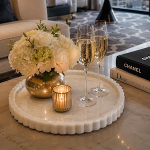 Könnte beinhalten: Ein rundes, weißes Marmortablett mit gewelltem Rand enthält eine goldene Vase mit weißen Hortensien und Rosen, einen kleinen goldenen Kerzenhalter mit brennender Kerze sowie zwei hohe Champagnergläser mit goldener Flüssigkeit neben Chanel- und Dior-Büchern.