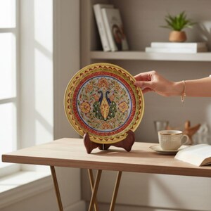 May include: A decorative plate with a peacock design. The plate features a yellow border, a red and blue inner ring, and a central image of two peacocks. The plate is displayed on a wooden stand on a light brown table.