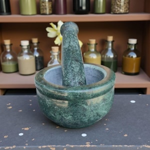 May include: A green marble mortar and pestle set. The mortar is a bowl shape with a matching pestle. The set is on a dark surface, with small glass bottles on a shelf in the background.