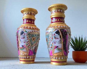 Könnte beinhalten: Zwei dekorative Vasen mit weißer Basis, verziert mit goldenen, roten und rosa Akzenten. Jede Vase zeigt ein gemaltes Porträt einer Frau in traditioneller Kleidung. Die Vasen haben eine bauchige Form und einen goldenen Rand.