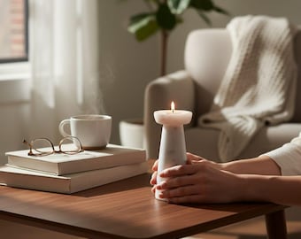 Op de afbeelding: Een brandende kaars in een witte en grijze marmeren kandelaar. De kandelaar staat op een donkerbruine houten tafel, met een kop koffie, boeken en een bril. Een gezellige woonkamersetting.
