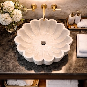 May include: A white, flower-shaped marble sink with a central drain, set on a dark countertop. Gold fixtures include a faucet and two handles. A bouquet of white flowers sits in a vase to the left. White towels and soap dispensers are also visible.