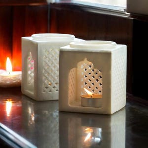 Puede incluir: Dos candelabros cuadrados blancos con intrincados diseños calados. Cada uno tiene una abertura circular en la parte superior y una abertura rectangular en el lateral. Una vela de té encendida está dentro de uno, emitiendo un brillo cálido.