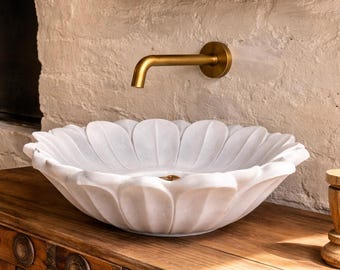 May include: A white marble sink with a petal design sits on a wooden cabinet. A gold faucet is mounted on the wall above the sink. The sink is a unique, decorative bathroom fixture.