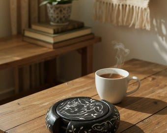 Può includere: Un set di sottobicchieri neri con un motivo floreale e una tazza da tè bianca su un tavolo di legno. Una piccola pianta in vaso e libri sono su un tavolo vicino. Un arazzo decorativo è sullo sfondo.