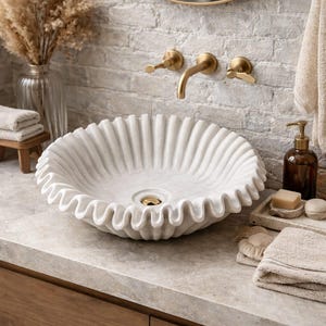 May include: A white, ruffled-edge marble sink with a gold drain and faucet. The sink is set on a light-colored countertop with a wooden cabinet below. A decorative arrangement of dried flowers and towels complete the bathroom scene.