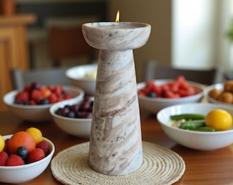 Op de afbeelding: Een marmeren kandelaar met een brandende kaars, met een beige en bruin wervelpatroon. De kandelaar staat op een ronde, geweven placemat. Schalen met fruit omringen de kandelaar op een houten tafel.