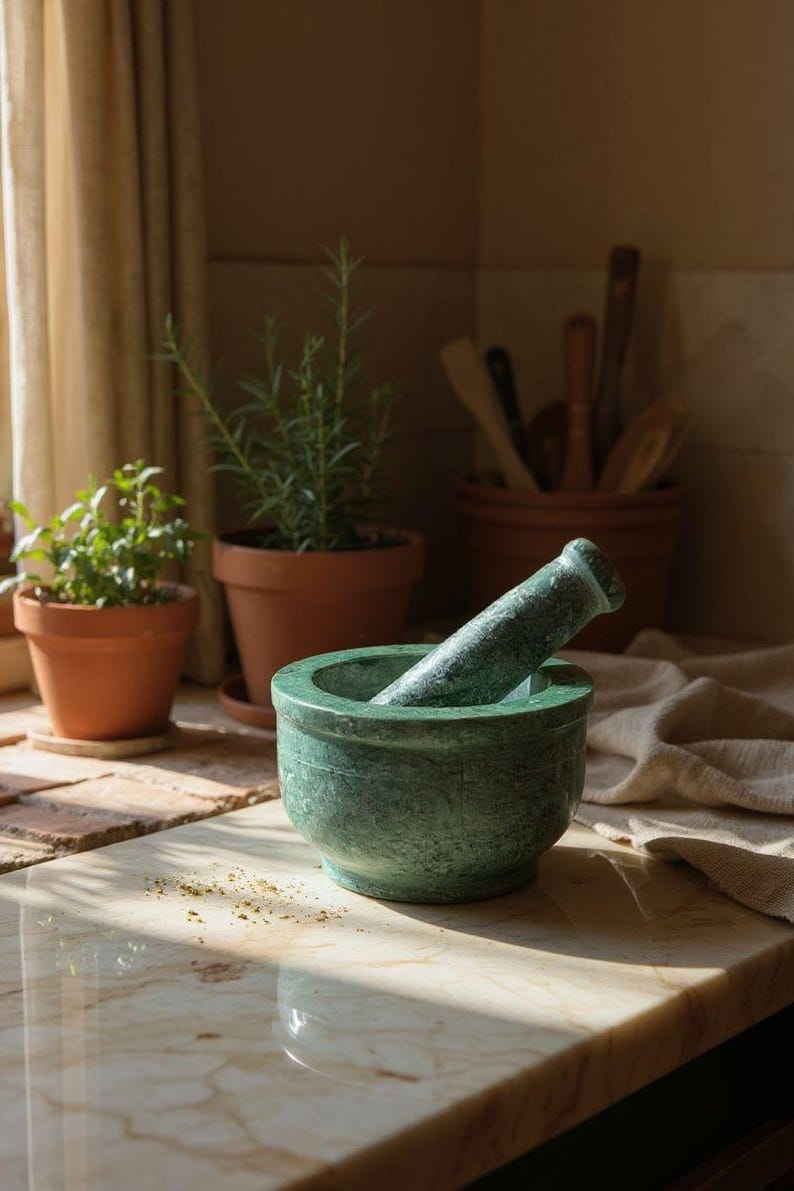 May include: A green marble mortar and pestle set. The mortar is round with a matching pestle resting inside. The set is on a marble countertop, with potted herbs and wooden utensils in the background.