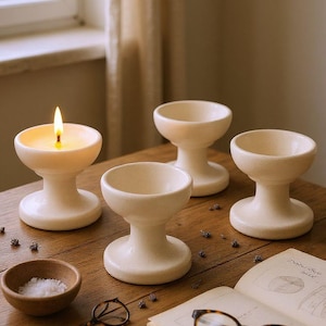 May include: Four off-white ceramic candle holders on a wooden table, one lit with a flame. A small wooden bowl with salt, a book, and eyeglasses are also on the table. The candle holders have a pedestal base and a shallow bowl shape.