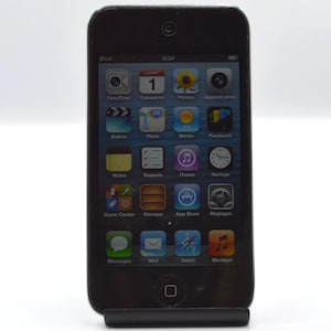May include: Black iPod touch with a touch screen displaying various app icons, including FaceTime, Calendar, and App Store. The device is held upright in a black stand. The time displayed on the screen is 0:34.