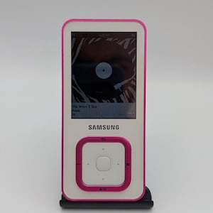 May include: White Samsung MP3 player with a pink trim and details. The screen shows a music album cover and song details. The player features a square control pad with a central button and a Samsung logo.