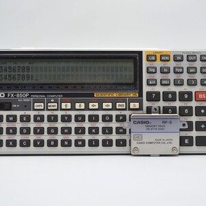 May include: A Casio FX-850P personal computer with a keyboard and display screen. The calculator has a silver and black color scheme, with a memory pack labeled "RP-8" next to it. The screen displays numbers.