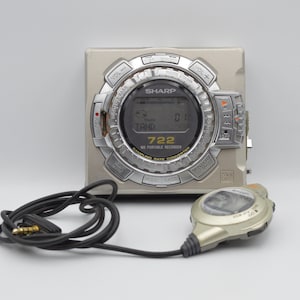 May include: A silver and grey Sharp MD portable recorder with a circular display and multiple buttons. The recorder has a matching remote control and a black cord with a gold-coloured plug. The display shows the text "TAND" and "722".