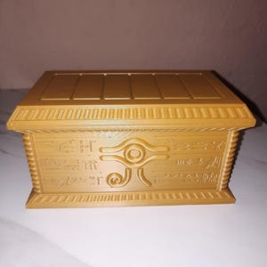 Könnte beinhalten: Eine goldfarbene Kartenbox mit einem ägyptischen Design, das Hieroglyphen und das Auge des Horus zeigt.
