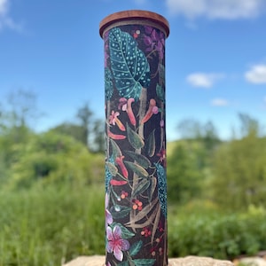 May include: A tall, cylindrical decorative object with a wooden top and base. The body is covered in a dark fabric with a colorful botanical print featuring leaves, flowers, and berries. The object is set against a natural outdoor backdrop.